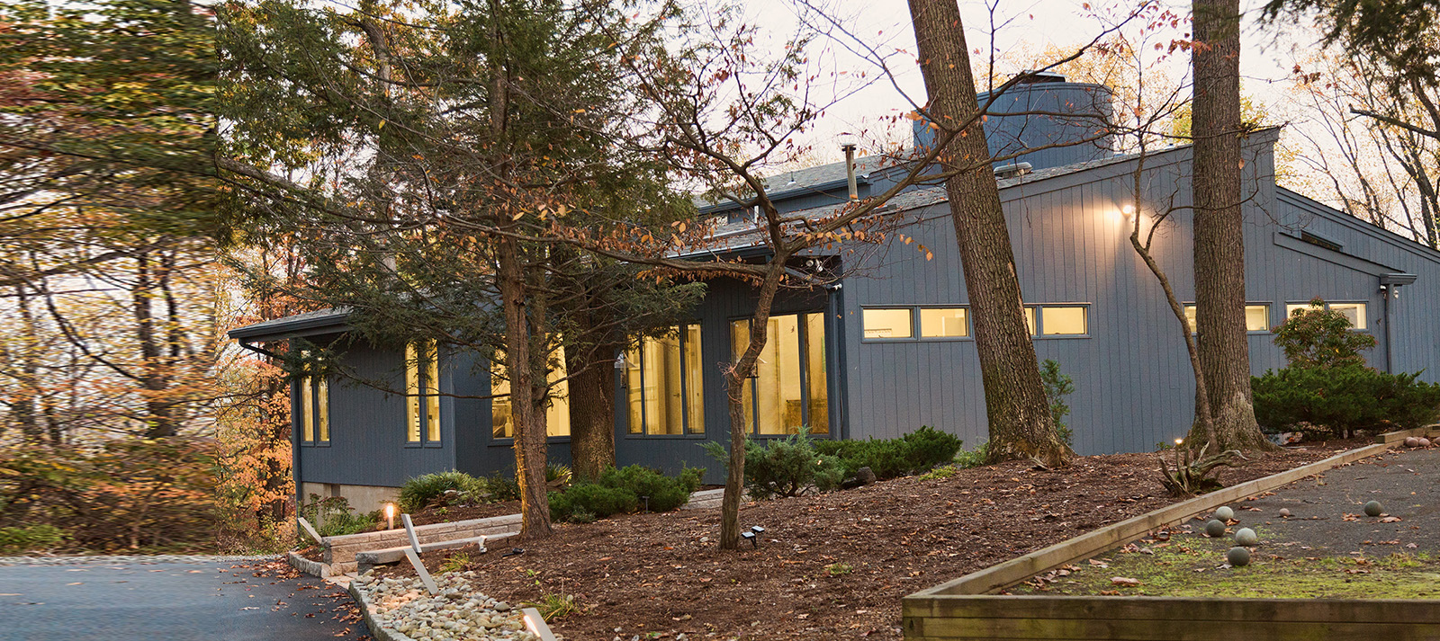 The image shows a large, modern two-story building with a prominent front entrance and a covered porch. The architecture features clean lines and a mix of dark and light exterior finishes. There is a clear sky above the building, suggesting it may be late afternoon or early evening.