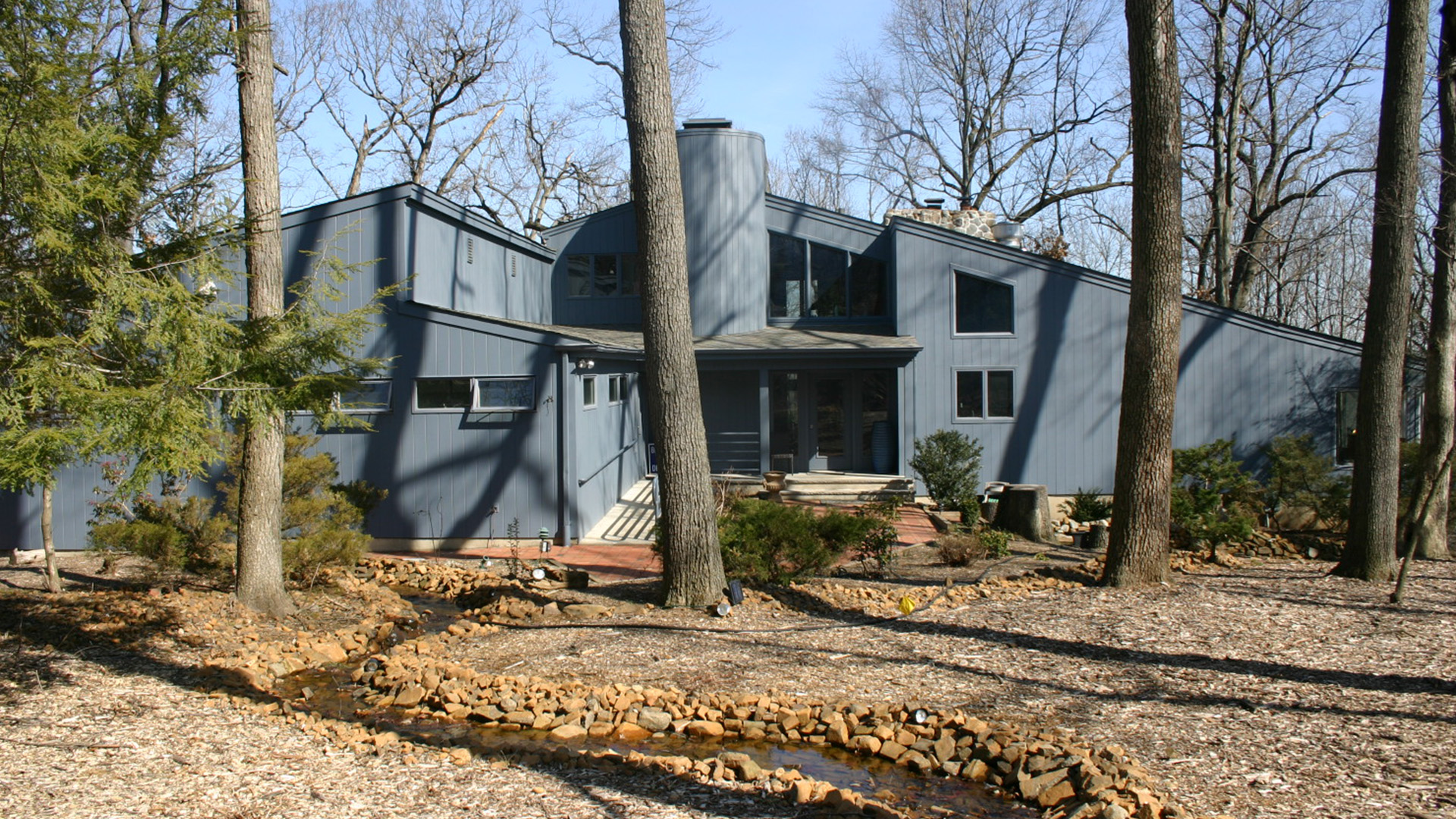 The image depicts a two-story house with a prominent blue exterior, set in a wooded area with trees and a path leading up to the house.